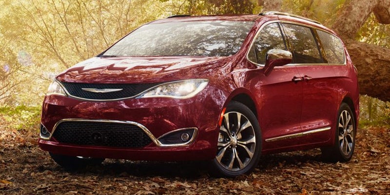 A shiny maroon minivan parked on a fallen leaves covered forest floor, against sunlit autumn trees.