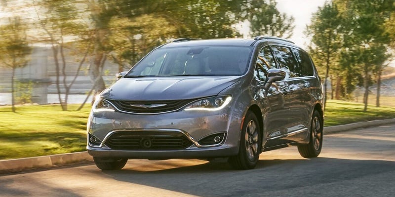 A silver minivan drives on a paved road, blurred trees and a distant bridge in gold light.