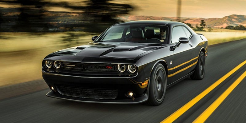 A black Dodge Challenger with yellow racing stripes drives along a highway with a blurred background.