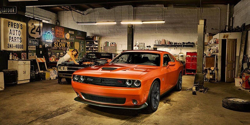 An orange muscle car in a garage with a woman leaning over a black classic car, surrounded by car signs.