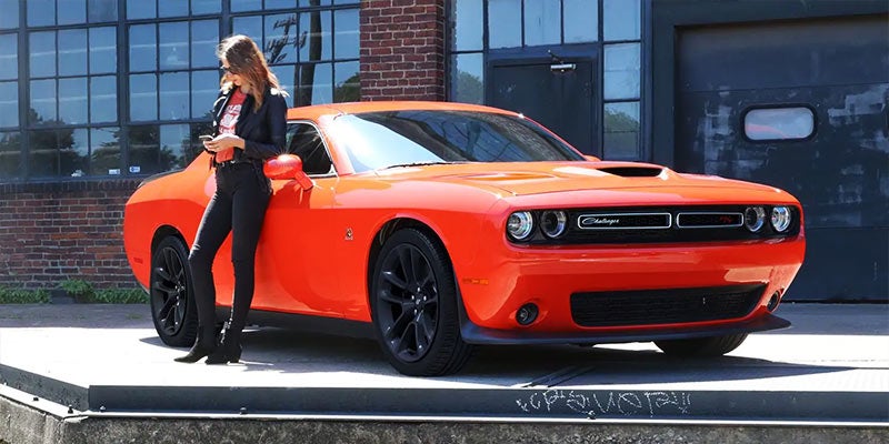 Woman in black leans on a bright orange Dodge Challenger, looking at her smartphone in a brick alley