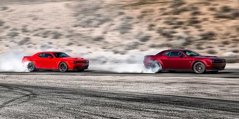 Two red Dodge Challenger cars doing burnouts, creating large clouds of smoke on an asphalt road.
