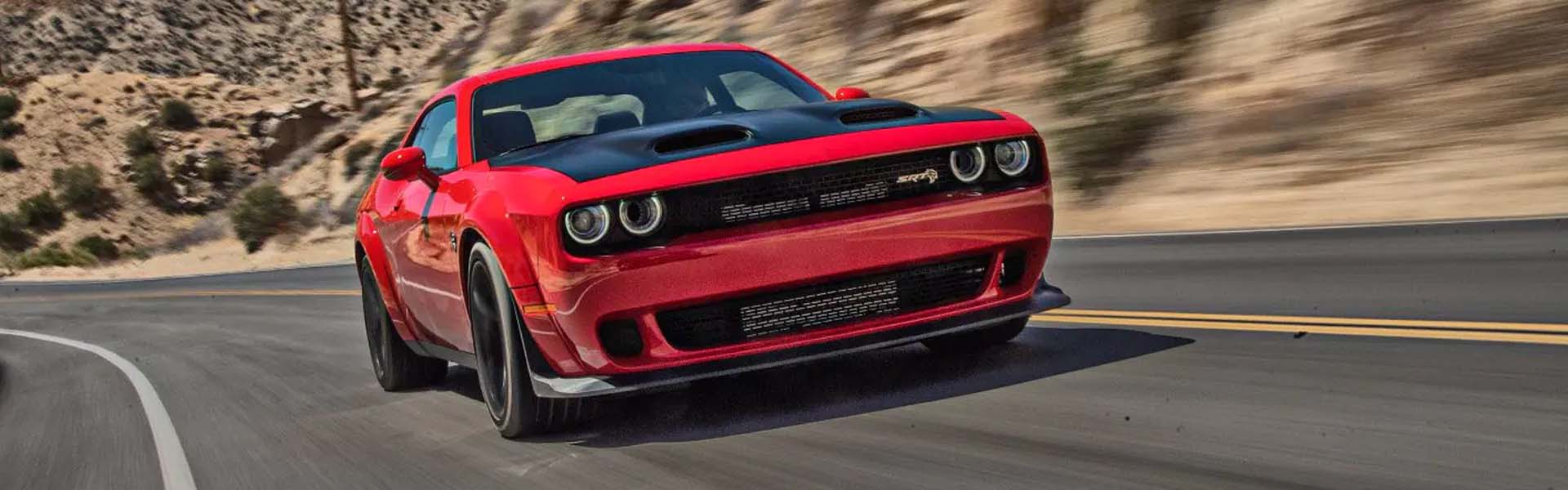A red Dodge Challenger SRT Hellcat with a black hood speeding on an asphalt road with blurry desert hills in the background.