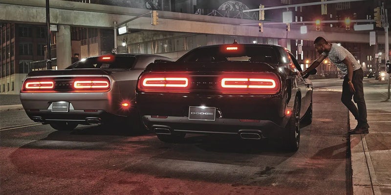 Two Dodge Challengers at night, one silver and one black, with illuminated taillights while a man leans on the black car.