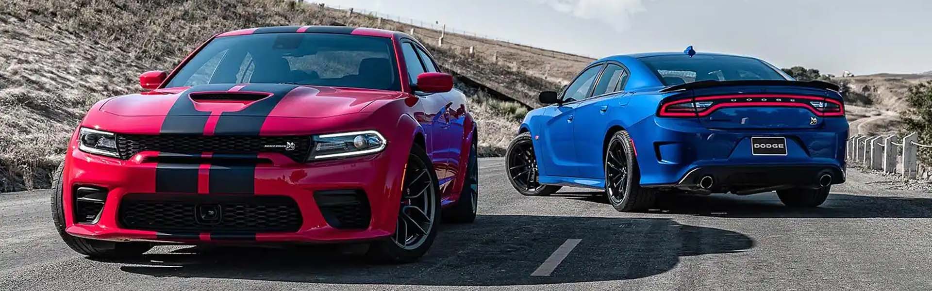 A red Dodge Charger with black racing stripes faces forward, and a blue Charger faces away on a paved road.