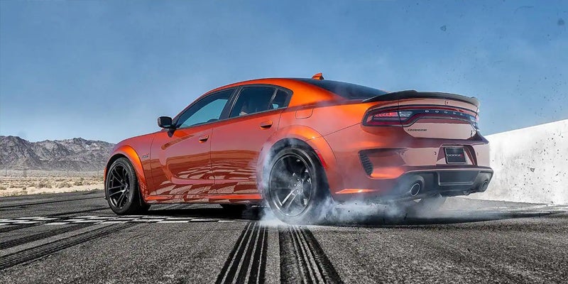 Rear 3/4 view of an orange Dodge Charger smoking its tires on a desert drag strip under a bright blue sky.
