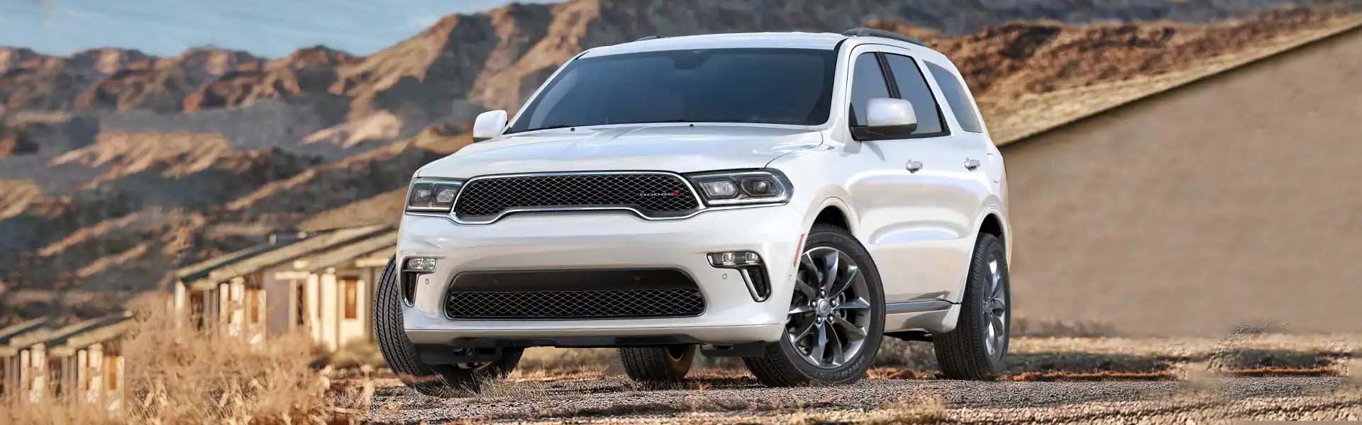 A white Dodge Durango, with a dark grille and headlights, parked on a dirt road in front of desert mountains.