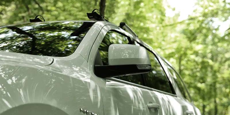 White truck with reflective windows and roof racks, parked among green trees, dappled with sunlight.