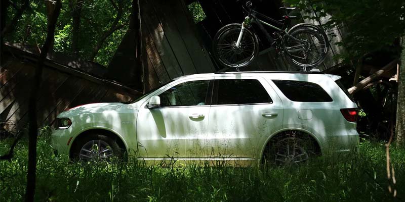 Mud-splashed white SUV with two bikes on the roof rack, parked in tall grass near a collapsed wooden structure.