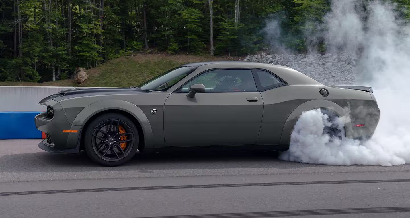 Gray Dodge Challenger creating a large smoke cloud from its spinning rear tire on a race track.