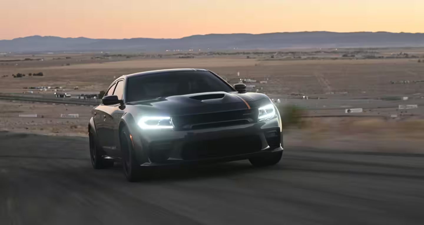 Dark gray Dodge Charger SRT Hellcat with bright headlights speeding along a road in a desert landscape at dusk.