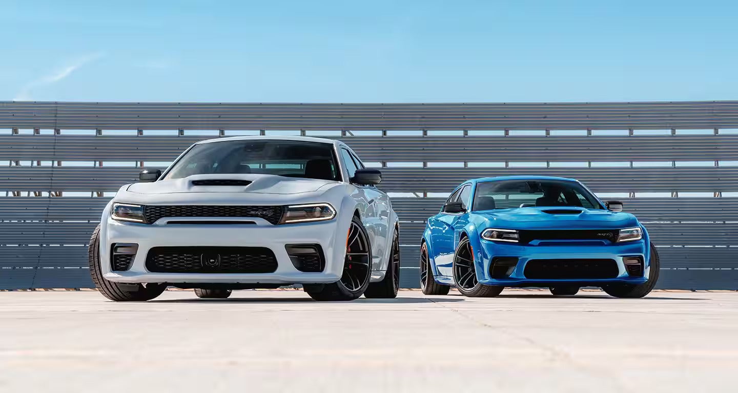 A white and a blue Dodge Charger SRT Hellcat parked on a concrete lot in front of a metal barrier.