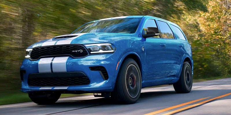 A blue Dodge Durango SRT with white racing stripes on an autumn road, speeding with trees blurring in the background.