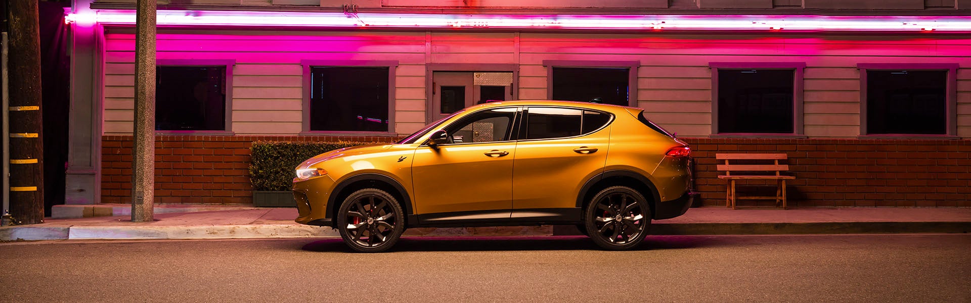 A golden Maserati Grecale SUV parked on a city street at night, with pink neon lights above.