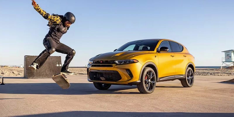 A skater in full gear jumps on a skateboard in front of a golden vehicle on a paved lot by a beach.