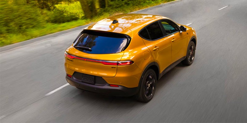 Rear view of a golden yellow Dodge Hornet R/T SUV driving on a road lined with green trees.