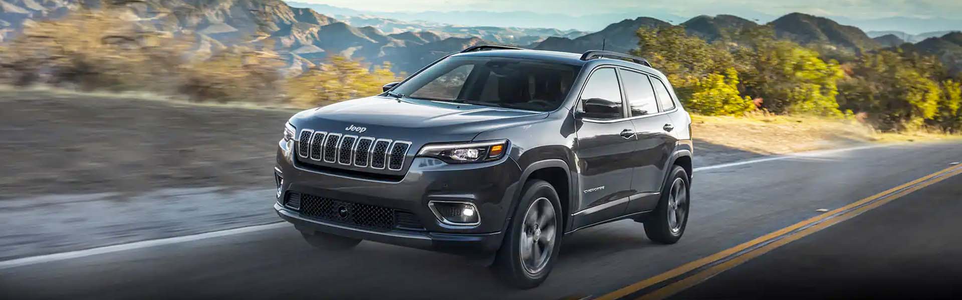 A dark gray Jeep Cherokee, angled towards the left, driving on a road with mountains in the background.