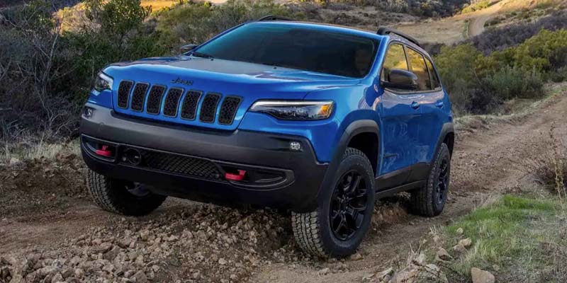 Blue Jeep Cherokee Trailhawk driving off-road over rocky terrain, with a dirt track and hills in the background.