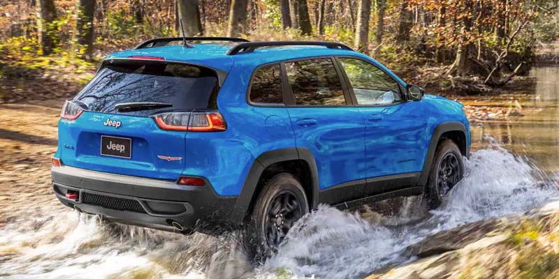 Blue Jeep Cherokee driving through a rocky stream, splashing water, in a wooded area.