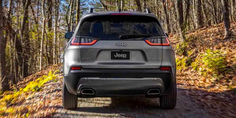 Rear view of a grey Jeep Cherokee driving on a dirt road through a forest with autumn foliage.