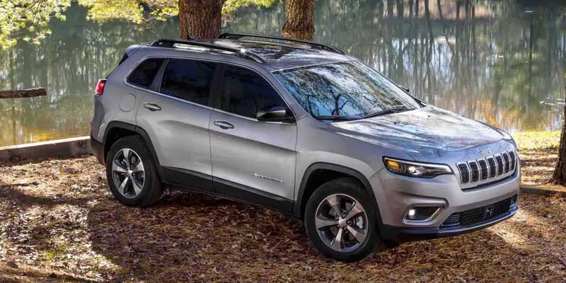 Silver Jeep Cherokee parked on brown fallen leaves next to a serene body of water reflecting trees.