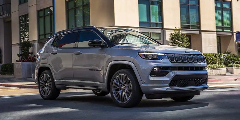 A silver Jeep Compass with a black roof and black wheels on an urban street with buildings in the background.
