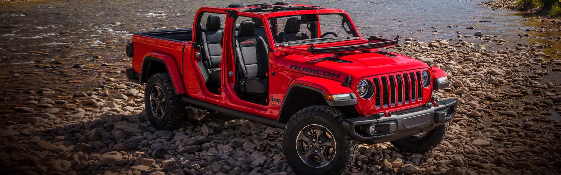A red Jeep Gladiator Rubicon pickup truck, doors and roof removed, parked on a rocky riverbank.