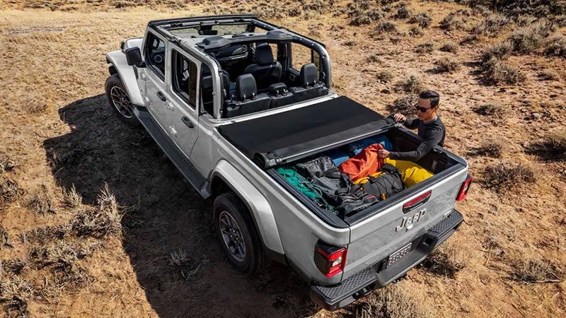 Overhead view of a silver Jeep Gladiator truck with its roof down. A man loads colorful camping gear into the bed.
