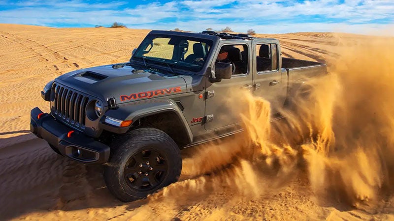 Gray Jeep Gladiator Mojave kicking up a large cloud of sand as it drives over desert dunes.
