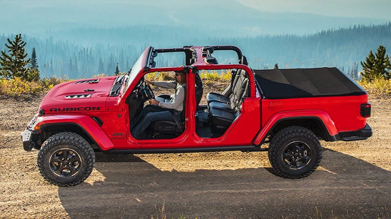 Red Jeep Gladiator Rubicon pickup truck, doors removed, parked on a dirt road in a mountainous forest.