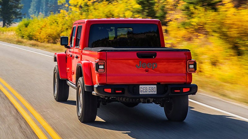 Rear view of a red Jeep Gladiator truck driving on a tree-lined road with yellow/orange fall foliage.