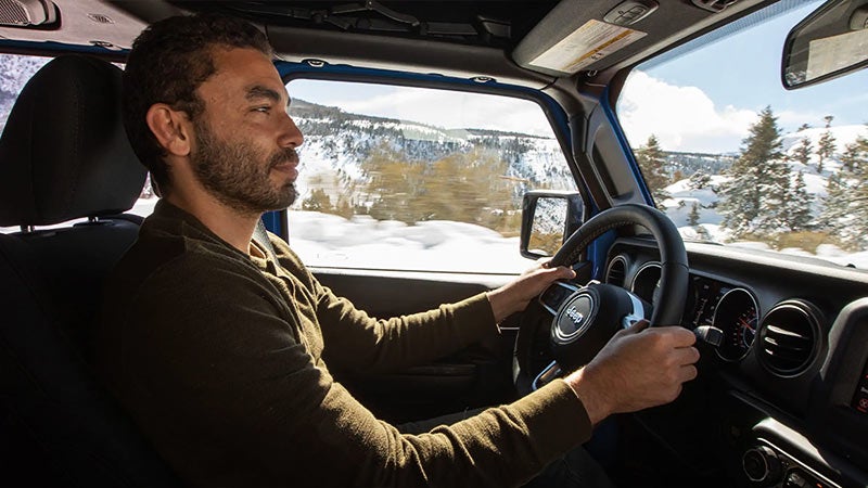 A man with a beard driving a Jeep in a snowy mountain landscape, hands on the wheel.