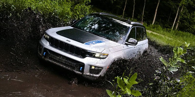 White Jeep Grand Cherokee 4xe splashing through deep mud, angled toward the viewer with jungle foliage.