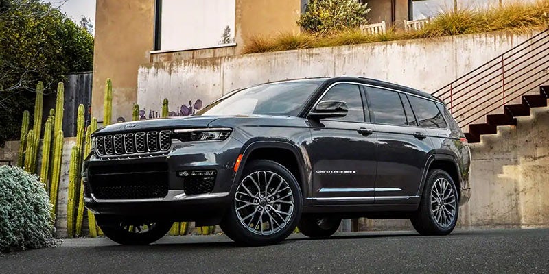 A dark gray Jeep Grand Cherokee parked beside a concrete wall with tall cacti and an outdoor staircase.
