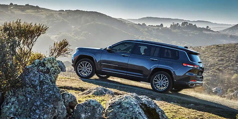 Dark gray SUV on a dusty mountain road, surrounded by rocks, trees, and rolling hills under a hazy sky.