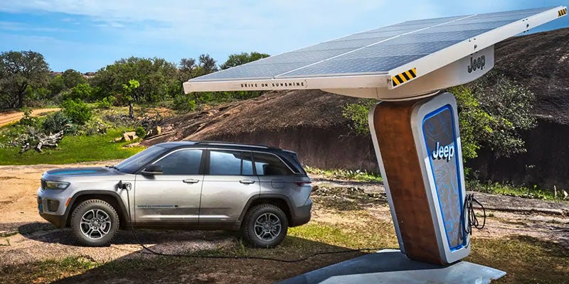A grey Jeep Grand Cherokee 4xe is charging at a solar-powered Jeep station in a rustic, rocky landscape.