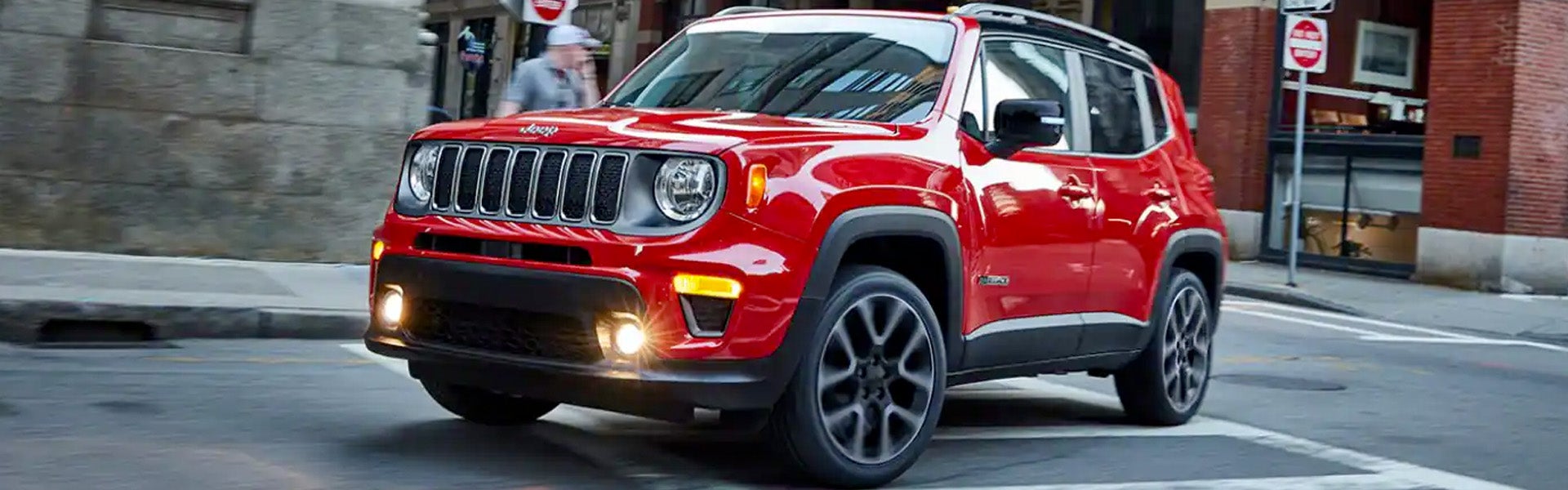 A shiny red Jeep Renegade SUV drives down a city street with its headlights on.