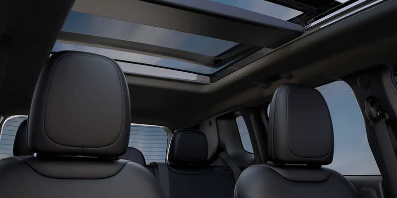 Black car interior with a panoramic sunroof open to the blue sky, viewed from the rear passenger seats.