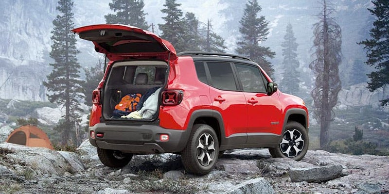 Red Jeep Renegade with open trunk full of camping gear in a foggy, rocky, and forested mountain landscape.
