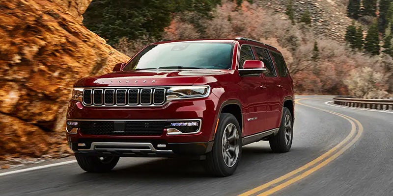 A red Wagoneer SUV drives on a winding road with mountains and trees in the background.
