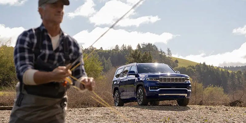 A man fly-fishing in a shallow river, with a blue Jeep Wagoneer parked behind him.