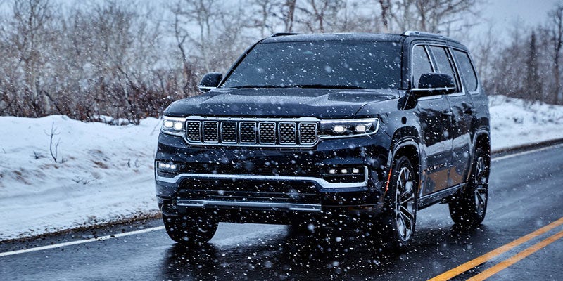 A black Jeep Grand Wagoneer driving on a wet, snowy road in a winter landscape with trees in the background.