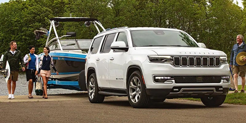 White Wagoneer SUV towing a blue boat, with a family of water skiers walking past the vehicle.