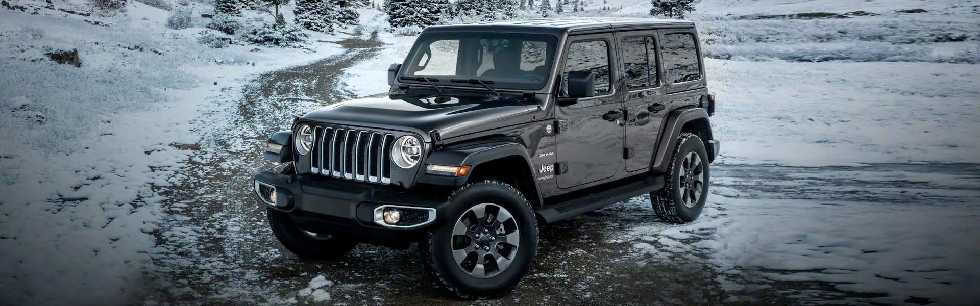 A dark gray Jeep Wrangler Unlimited, front left quarter view, parked on a snow-covered dirt road in wintry woods.