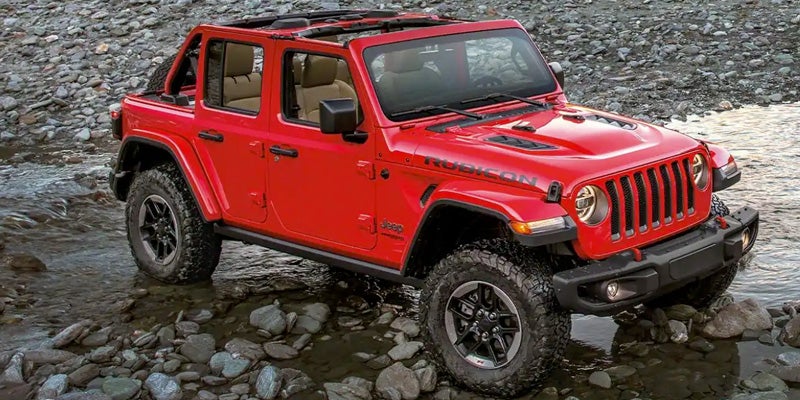 A red Jeep Wrangler Rubicon parked in shallow water on a rocky river bed.