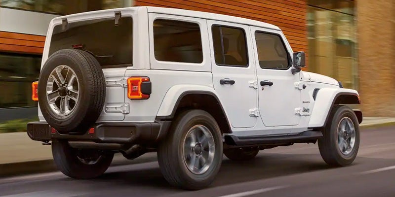 A white 4-door Jeep Wrangler Unlimited driving on a paved road, viewed from the rear passenger side.