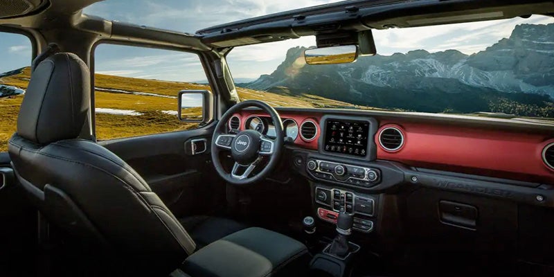 Interior of a Jeep Wrangler with red dashboard accents, an open roof, and a mountain landscape visible through the windows.