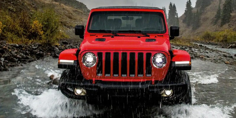 A red Jeep Wrangler drives through a rocky river, with splashing water around its tires, towards the viewer.