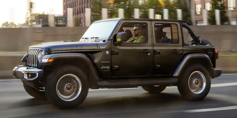 Dark grey Jeep Wrangler Unlimited driving on a highway with city buildings in the blurred background.