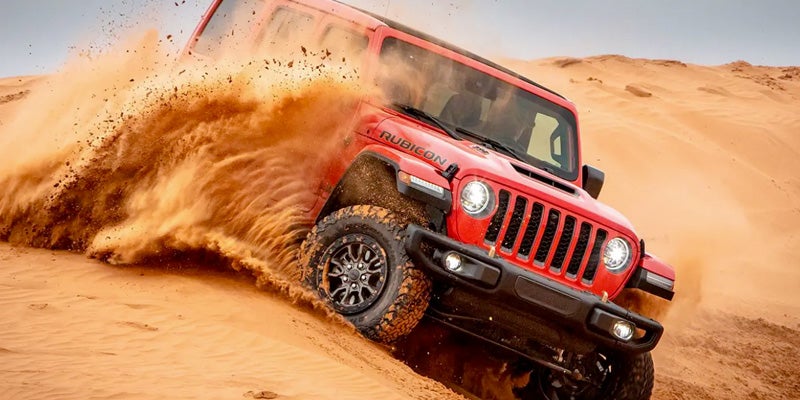 A red Jeep Rubicon, kicking up a large spray of sand, drives angled down a steep desert dune.